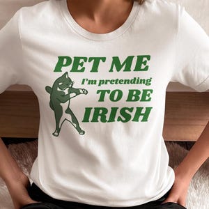 May include: White t-shirt with a green graphic of a cat and the text "PET ME I'm pretending TO BE IRISH". The cat is depicted in a dancing pose. The text is in a bold, green font.