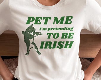以下が含まれることがあります： 白いTシャツに、緑色の猫のグラフィックと「PET ME I'm pretending TO BE IRISH」の文字。猫は踊るポーズで描かれています。テキストは太字の緑色です。