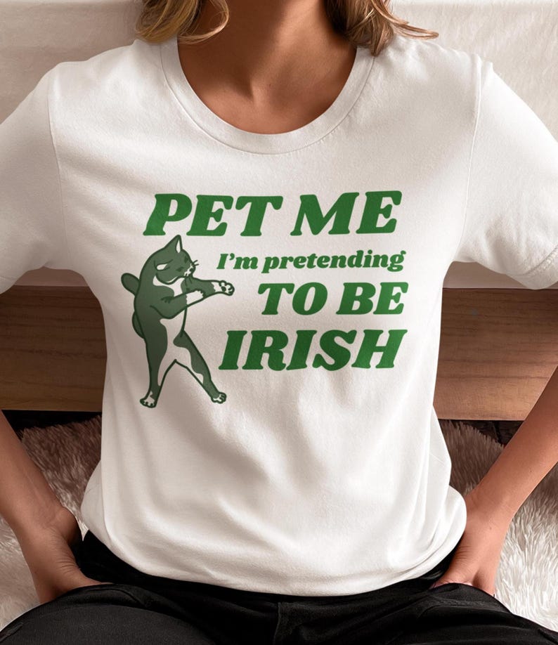 May include: White t-shirt with a green graphic of a cat and the text "PET ME I'm pretending TO BE IRISH". The cat is depicted in a dancing pose. The text is in a bold, green font.