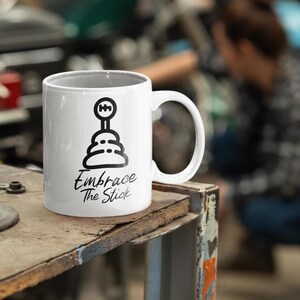 May include: White ceramic mug with a black illustration of a stick shift and the text "Embrace The Stick".