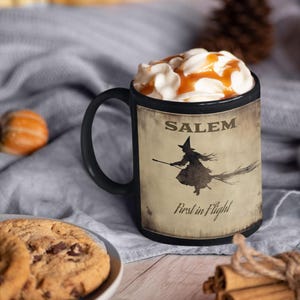 May include: Black ceramic mug with a white and brown design. The mug features a silhouette of a witch flying on a broomstick with the text "Salem First in Flight".