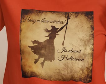 May include: Orange t-shirt with a vintage-style graphic of a witch flying on a broomstick. The graphic has the text "Hang in there witches!" and "It's almost Halloween".
