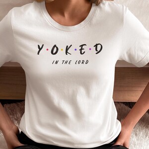May include: White t-shirt with the text "Y.O.K.E.D" in black, with the letters O, K, and E in different colors. Below the text is the phrase "IN THE LORD" in black. The shirt is made of a soft material.