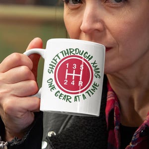Könnte beinhalten: Weiße Keramik-Tasse mit einer rot-weißen Grafik eines Autoschalthebels, mit dem Text "SHIFT THROUGH XMAS ONE GEAR AT A TIME" in Grün und Weiß. Die Tasse wird von einer Person gehalten.