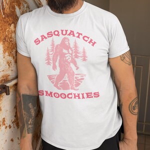 Peut inclure: T-shirt blanc avec un graphique rose d'un sasquatch marchant dans une for&ecirc;t avec le texte "Sasquatch Smoochies".