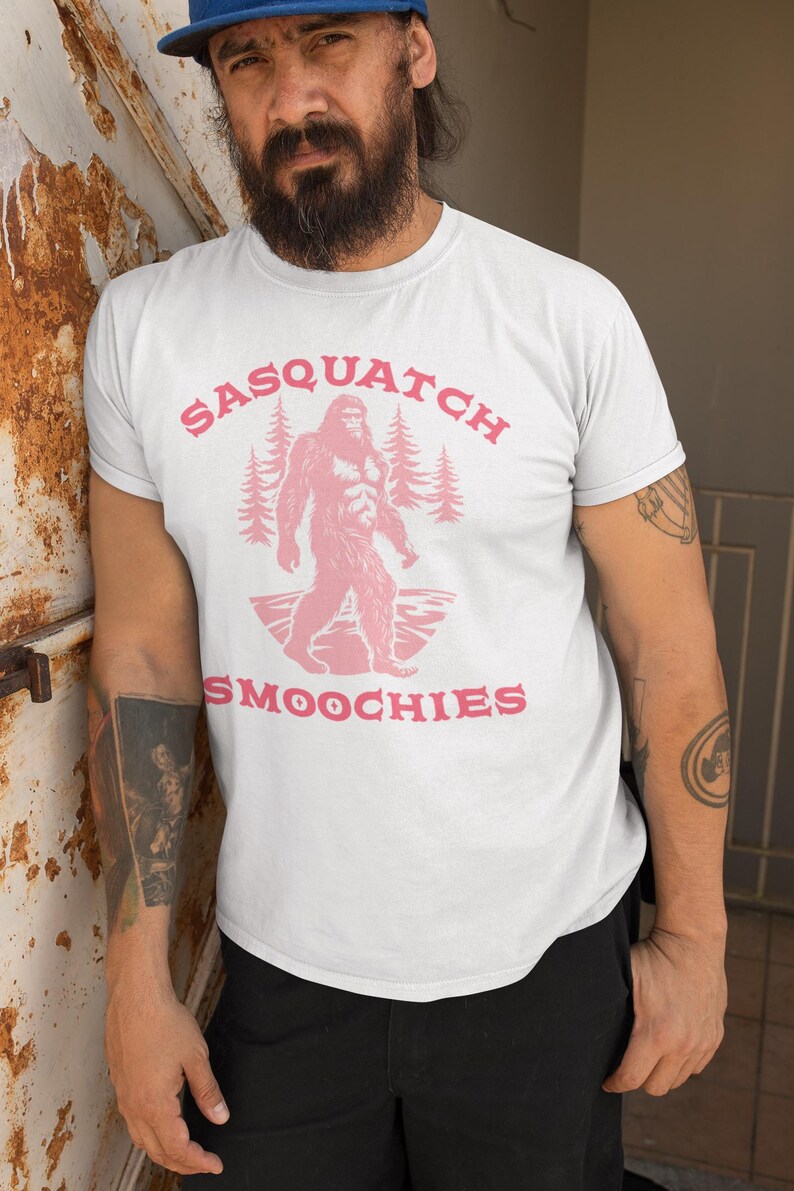 Peut inclure: T-shirt blanc avec un graphique rose d'un sasquatch marchant dans une for&ecirc;t avec le texte "Sasquatch Smoochies".