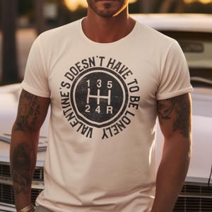 May include: A white t-shirt featuring a black graphic of a car's gear shift pattern. The text surrounding the graphic reads "Valentine's Doesn't Have To Be Lonely".