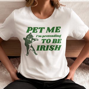 May include: White t-shirt with green text and a cartoon cat graphic. The text reads "PET ME I'm pretending TO BE IRISH". The cat is depicted in a playful pose. The shirt is a crew neck style.