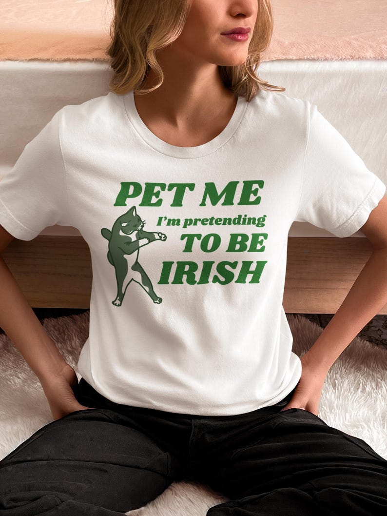 May include: White t-shirt with green text and a cartoon cat graphic. The text reads "PET ME I'm pretending TO BE IRISH". The cat is depicted in a playful pose. The shirt is a crew neck style.
