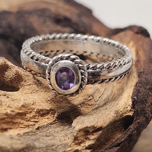 May include: A silver ring featuring a central oval-shaped purple gemstone. The band is constructed with a twisted rope-like texture that encircles the entire circumference, including a matching twisted metal border surrounding the purple stone.
