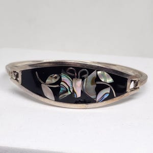 Puede incluir: Pulsera plateada con un centro de esmalte negro que presenta un diseño de mariposa y hojas. La mariposa está hecha de incrustaciones iridiscentes y nácar. La pulsera tiene un cierre de bisagra.