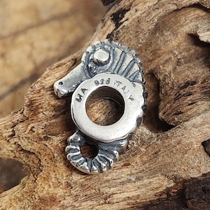 Puede incluir: Un charm detallado de plata con forma de caballito de mar, con un gran agujero central. El charm está marcado con "MA 925 ITALY". El caballito de mar tiene detalles texturizados y una cola curvada.
