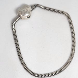 Puede incluir: Pulsera plateada con cierre en forma de corazón grabado con la palabra "MEMORIES". La pulsera tiene un diseño de cadena de serpiente.