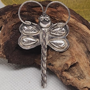 May include: A silver dragonfly pendant with intricate detailing. The pendant features a braided body and detailed wings. The wings have a floral design. The pendant is suspended from a silver chain. The dragonfly is a symbol of transformation.