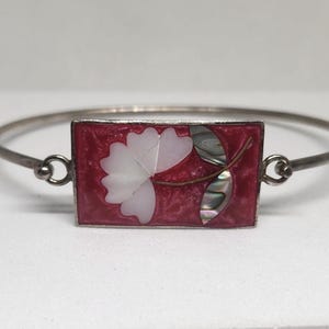 May include: A silver-toned bangle bracelet with a rectangular centerpiece. The centerpiece features a red background with a white flower and iridescent accents. The bracelet is a delicate piece of jewelry.