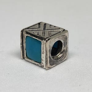 May include: A silver-toned cube-shaped charm with a turquoise blue square inlay. The charm has a geometric design on the top and is designed to be strung on a bracelet or necklace. The charm is made of metal.