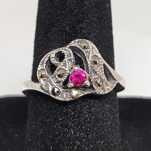 May include: A silver ring with a heart-shaped design, featuring a vibrant pink gemstone at its center. The ring is adorned with small, round, gold-colored accents and is displayed on a black surface.
