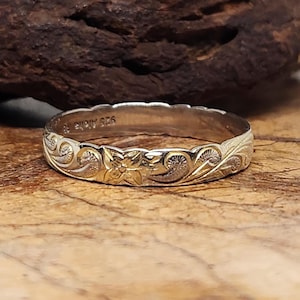 Puede incluir: Un anillo de plata con un diseño floral y de volutas. La banda del anillo presenta un patrón repetido de flores y hojas. Está colocado sobre un fondo de madera. El anillo está marcado con "925 Aloha 18".