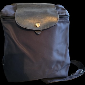 Puede incluir: Una mochila de nailon azul oscuro con una solapa de cuero negro y una hebilla plateada. La solapa tiene la palabra "Longchamp" grabada en relieve.