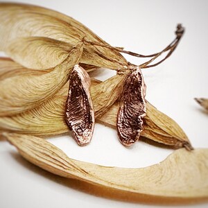 May include: A pair of rose gold earrings shaped like maple seeds. The earrings are delicate and have a natural, organic look.