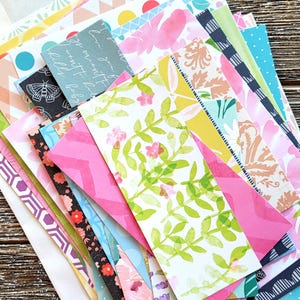May include: A collection of colorful patterned paper sheets in a white envelope. The papers feature various designs, including floral, geometric, and abstract patterns in shades of pink, green, blue, and yellow. The papers are arranged on a wooden surface.