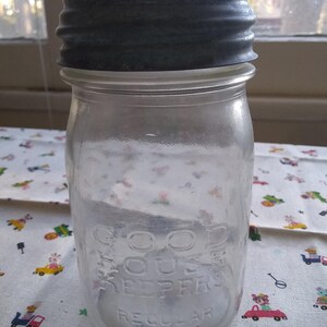 May include: A clear glass jar with a dark gray metal lid. The jar is labeled "GOOD HOUSE KEEPERS REGULAR MASON". The jar is sitting on a patterned fabric.