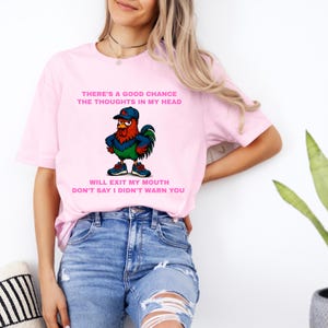 May include: Light pink t-shirt with a cartoon rooster graphic wearing a baseball cap and sneakers. Text on the shirt reads: "THERE'S A GOOD CHANCE THE THOUGHTS IN MY HEAD WILL EXIT MY MOUTH DON'T SAY I DIDN'T WARN YOU."