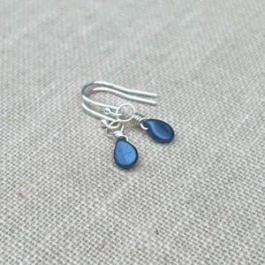 May include: Silver drop earrings featuring dark blue, teardrop-shaped beads. The earrings have a simple hook design and are presented on a neutral, textured surface. The beads have a subtle shimmer, reflecting light.