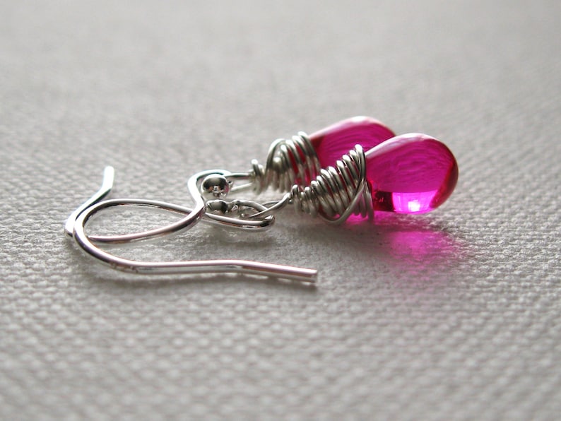 On a white canvas rests a pair of teardrop bead earrings.  The clear glass pear shaped teardrop bead is a fun shade of hot pink. Wrapped around the top half of the beads is sterling silver wire, which at the small end loops to attach to ear wires.