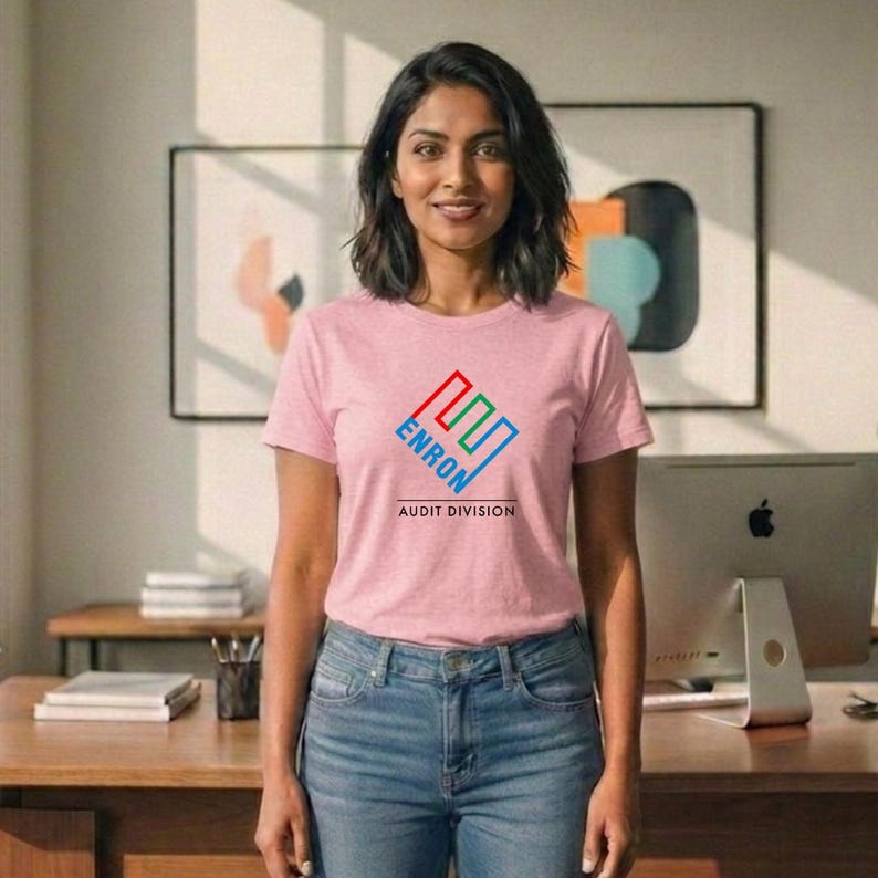 May include: A light pink t-shirt with the word "ENRON" in a blue, red, and green geometric design. Below the design, the text "AUDIT DIVISION" is printed in black. The person is wearing blue jeans.