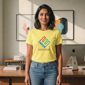 May include: A yellow t-shirt with the text "ENRON AUDIT DIVISION" in a colorful logo. The shirt is paired with blue jeans. The image is taken indoors, with a desk and computer visible in the background.