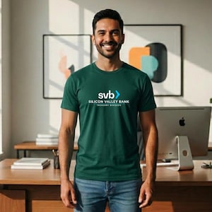 May include: A smiling person wearing a dark green crew neck t-shirt with the text "svb" and "SILICON VALLEY BANK TREASURY DIVISION" in white. The person is standing in front of a wooden desk with a computer and artwork.