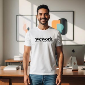 May include: White crew neck t-shirt with the black text "wework STRATEGY DEPARTMENT". The person is wearing blue jeans. The background includes a desk, computer, and framed artwork.