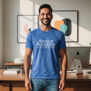 Op de afbeelding: Een blauw T-shirt met ronde hals en de witte tekst "ARTHUR ANDERSEN ETHICS COMMITTEE". Het shirt heeft een getextureerde uitstraling. De persoon draagt een blauwe spijkerbroek.