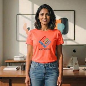May include: An orange t-shirt with the text "ENRON AUDIT DIVISION" in blue, green, and red. The person is wearing blue jeans. A computer monitor and artwork are in the background.