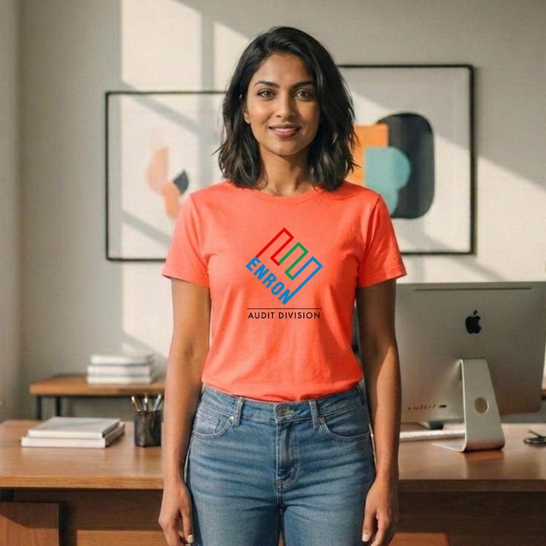 May include: An orange t-shirt with the text "ENRON AUDIT DIVISION" in blue, green, and red. The person is wearing blue jeans. A computer monitor and artwork are in the background.