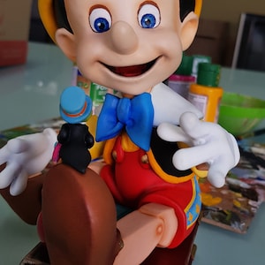 Könnte beinhalten: Eine Pinocchio-Figur, die auf einem geschlossenen Buch sitzt. Er trägt einen gelben Hut, eine blaue Fliege und ein rot-gelbes Outfit. In seiner linken Hand hält er eine kleine schwarze Figur.