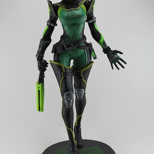 May include: A green and black action figure of a female character holding a weapon. The figure is posed in a dynamic stance with one leg forward and the other leg back. The figure is wearing a green and black suit with a belt and a weapon holster.