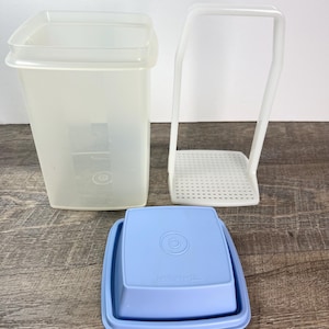 May include: A collection of Tupperware containers and accessories. Includes a tall, rectangular, translucent container, a white plastic rack, and a light blue square container with a lid. The containers are made of plastic and are designed for food storage.