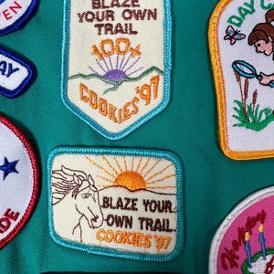 May include: Assortment of embroidered patches with various designs and text. One patch reads "BLAZE YOUR OWN TRAIL COOKIES '97" with a horse and sun graphic. Other patches include text like "ADVENTURE" and "DAY CAMP".