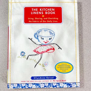 May include: A white book titled "THE KITCHEN LINENS BOOK" with a red rectangular banner. The cover features an embroidered teacup character and the text "Using, Sharing, and Cherishing the Fabrics of Our Daily Lives". Includes a Butterick transfer pattern for a vintage kitchen towel motif.