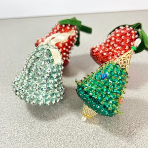 May include: Four decorative Christmas ornaments. Two are red, strawberry-shaped ornaments with green fabric leaves. One is a light blue bell-shaped ornament. The fourth is a green Christmas tree ornament with gold accents.