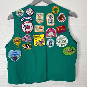 May include: A green Girl Scout vest adorned with various patches. Patches include "Memorial Day," "Girl Scouts on Parade," "Blaze Your Own Trail," and "Leader Daughter '98." The vest is hanging on a gray hanger.