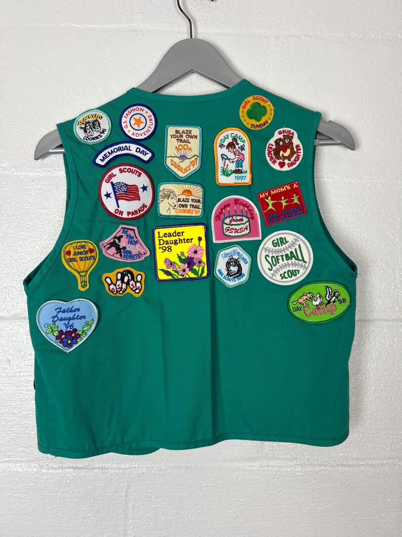 May include: A green Girl Scout vest adorned with various patches. Patches include "Memorial Day," "Girl Scouts on Parade," "Blaze Your Own Trail," and "Leader Daughter '98." The vest is hanging on a gray hanger.