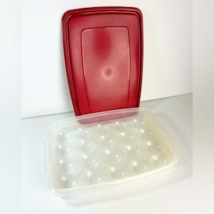 May include: A red rectangular lid sits above a clear plastic egg storage container. The container has a grid of rounded depressions to hold eggs. The lid is a solid, glossy red.