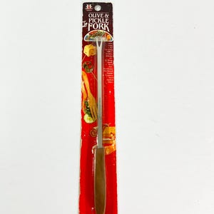 May include: An olive and pickle fork on a red and white cardboard backing. The fork has a stainless steel tine and a wooden handle. The packaging reads "OLIVE & PICKLE FORK".