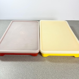 May include: Two rectangular food storage containers. One is light purple with a red base, and the other is yellow with a yellow base. Both have a lid and are on a gray surface.