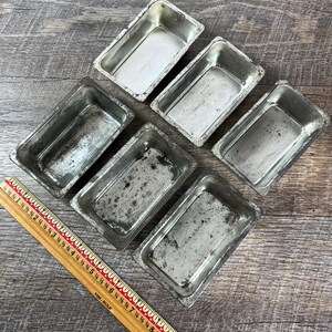 May include: Six small, rectangular, metal baking pans with a distressed, silver finish are arranged on a wooden surface. A wooden ruler with inch markings lies diagonally across the image, providing scale.