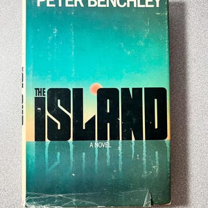 May include: Vintage paperback book titled "The Island" by Peter Benchley. The cover features a teal and orange gradient with the title in bold black letters. The book is a novel.