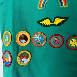 May include: Green fabric with various colorful embroidered patches. Patches include a rainbow, wings, and circular designs with images of a snowflake, a person, and other symbols. The fabric has a pocket.